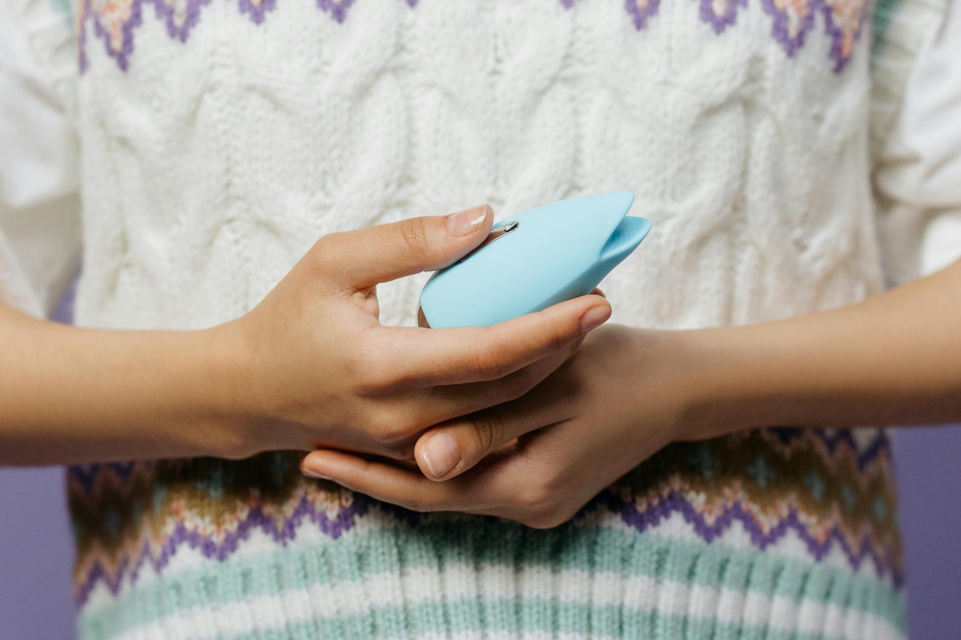 Close-up of hands holding a blue personal massager against a knitted sweater, suggesting comfort and self-care during vulnerability.